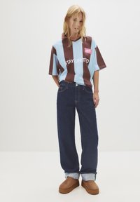 Polo tričko s oversized střihem v barevném bloku, modré a hnědé pruhy; obsahuje grafiku "Stay United". Spárované s volně střiženými tmavomodrými džíny a hnědými botami.