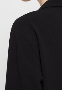 Černý blazer s vyztuženým límcem, hladkým materiálem a upravenými rameny, který se vyznačuje elegantním minimalistickým designem.