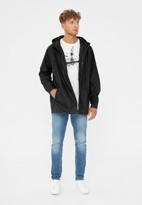 Giacca a vento nera con cappuccio, leggera e con chiusura a bottoni, indossata sopra una t-shirt bianca con grafica e abbinata a jeans azzurri e sneaker bianche.