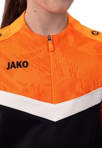 Maglietta sportiva con zip arancione e nera, caratterizzata da un motivo geometrico, una striscia bianca, una chiusura con zip e un logo sul petto.