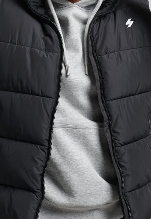 Persona que lleva una sudadera gris con cordones debajo de una chaqueta de plumas negra parcialmente subida, con un pequeño logo blanco en el pecho.