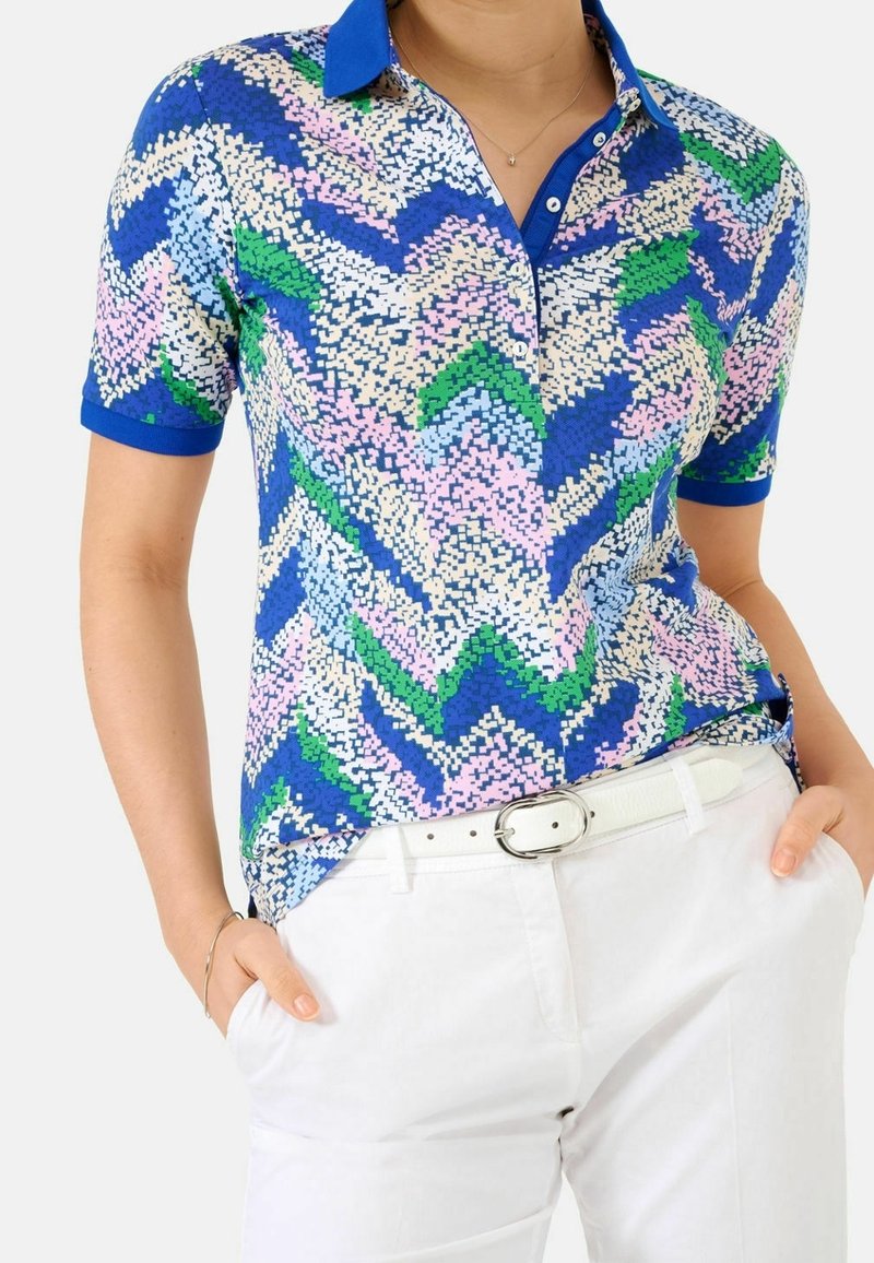 Femme portant un polo à manches courtes à motif zigzag coloré avec col bleu, ceinture blanche et pantalon blanc, mains dans les poches.