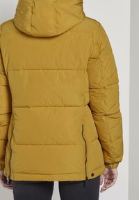 Hořčičně žlutá péřová bunda s kapucí, s prošívaným vzorem a bočními zipovými kapsami, s elastickými manžetami pro pohodlné nošení.
