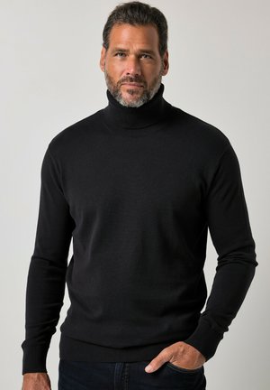 Mand med skæg iført sort rullekrave og mørke bukser, der står mod en ensfarvet lys grå baggrund med den ene hånd i lommen.