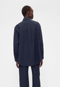 Camicia blu navy con bottoni realizzata in tessuto strutturato, con piega posteriore, maniche lunghe e orlo curvo.