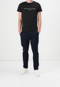 Mies, joka on pukeutunut mustaan Tommy Hilfiger -t-paitaan, tummansinisiin housuihin ja valkoisiin tennareihin, seisoo kädet taskuissa valkoista taustaa vasten.