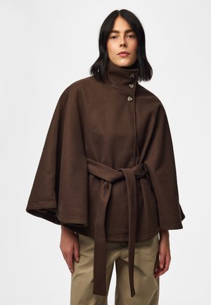 Femme portant un manteau cape marron foncé ceinturé avec un col montant et des boutons, associé à un pantalon beige sur un fond uni.
