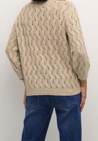 Pull beige en tricot avec un motif ondulé, manches longues et ourlet côtelé, associé à un jean en denim bleu. Présente des coutures ouvertes pour la ventilation.