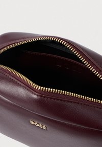 Βουργουνδί δερμάτινη θήκη με χρυσό φερμουάρ και λεία υφή; διαθέτει μαύρη εσωτερική επένδυση και ανάγλυφο λογότυπο μπροστά.