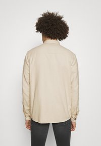Beżowa koszula z długim rękawem, z kołnierzykiem, prostym kroju i zapięciem na guziki przy mankietach, wykonana z teksturowanej tkaniny. Szew środkowy przebiega wzdłuż pleców.