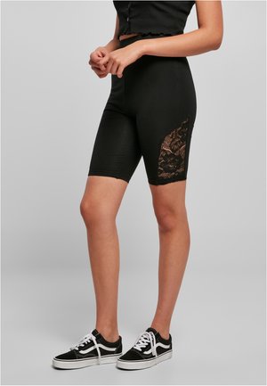 Kvinde iført sorte højtaljede shorts med blonder i siderne, sort crop top og sorte og hvide sneakers, der står mod en grå baggrund.