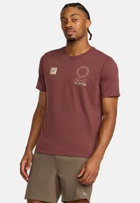 Bordowy bawełniany t-shirt z krótkimi rękawami, okrągłym dekoltem i grafiką z przodu, w tym logo oraz okrągłym detalem wzorniczym.