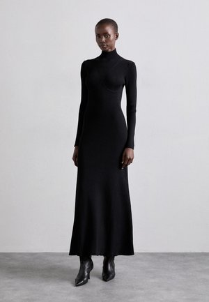 Rochie neagră cu guler înalt, cu fir ribbed, mâneci lungi, cu un corset ajustat și o fustă evazată. Purtați-o cu botine cu vârf ascuțit pe un fundal neutru.