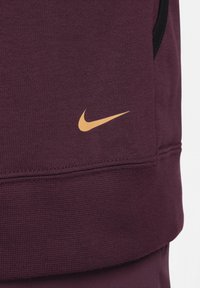 Sweat-shirt Nike bordeaux en tissu texturé, avec des accents noirs et un logo swoosh doré en bas à gauche.
