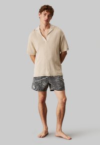 Polo de punto ribeteado beige de manga corta con cuello en V y collar ligero, combinado con shorts negros que presentan patrones ondulados blancos. Ajuste casual.