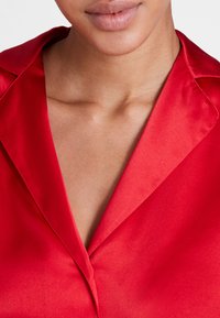 Blouse en satin rouge avec un col cranté et une texture lisse, présentant un léger brillant et des coutures nettes le long des bords.