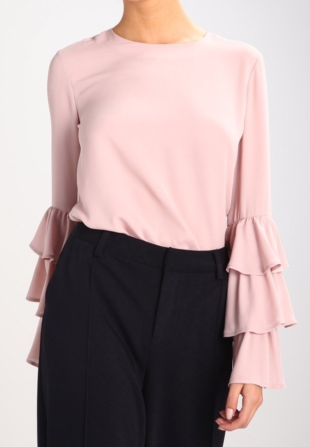 Blusa a maniche lunghe in morbido tessuto rosa presenta un colletto rotondo e maniche a strati con volant. Abbinata a pantaloni scuri sartoriali.