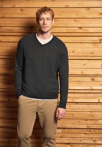Blonder Mann, der gegen eine Holzplankenwand steht, trägt einen dunkelgrauen V-Ausschnitt-Pullover über einem weißen Hemd und beigen Hosen.