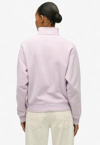 Kobieta z włosami upiętymi w kok, ubrana w jasnofioletowy sweter z golfem i jasnobeżowe spodnie, stojąca tyłem na jednolitym tle.