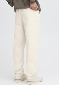 Light cream cotton pants with a relaxed fit and two back pockets, paired with white sneakers. Smooth texture and minimalist design.