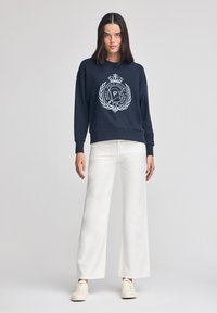 Felpa navy con logo bianco, vestibilità rilassata, collo rotondo e spalle scese. Abbinata a pantaloni ampi color crema e scarpe da ginnastica bianche.