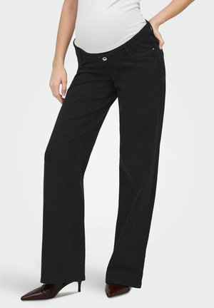 Personne enceinte portant un jean de maternité noir et des talons pointus marron, debout avec une main dans la poche sur un fond uni.