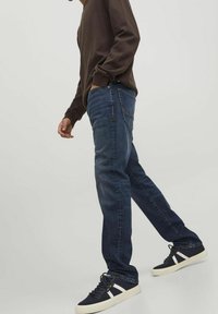 Jeans blu scuro a gamba dritta, abbinati a una camicia marrone a maniche lunghe e sneakers blu navy con dettagli bianchi.