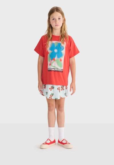 Jeune fille aux longs cheveux blonds portant un t-shirt rouge avec un motif de fleur bleue, une jupe blanche à imprimés de fruits orange, des chaussettes blanches et des chaussures rouges.