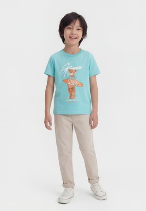 Lachende jongen staand, gekleed in een lichtblauw t-shirt met teddybeerafbeelding en de tekst "Guess", beige broek en witte sneakers tegen een witte achtergrond.
