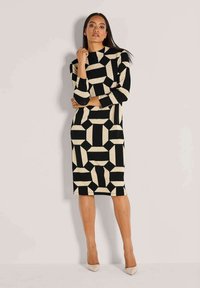 Frau, die gegen eine Wand steht, in einem knielangen, langärmeligen Kleid mit geometrischem Muster in Schwarz und Beige sowie beigen spitzen Schuhen.