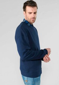 Felpa blu navy con scollatura a girocollo e polsini a costine, indossata sopra una camicia a quadri. Tessuto testurizzato, vestibilità rilassata, design minimalista.