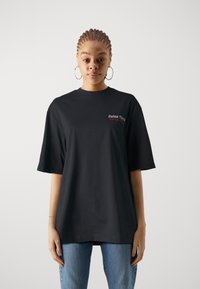 Sort bomulds t-shirt med korte ærmer, rund halsudskæring, med rød og hvid tekstlogo på brystet, parret med blå jeans.