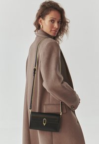 Borsa a tracolla in pelle nera con chiusura in metallo dorato, forma rettangolare e texture liscia, indossata sopra un cappotto marrone chiaro.