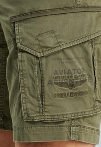 Olivenfarbene Cargo-Shorts aus leichtem Material. Verfügt über mehrere Taschen und bestickte Logos. Nähte entlang der Kanten sichtbar.