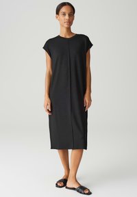 Zwarte midi-jurk met korte mouwen, relaxte pasvorm, slank ontwerp, zachte textuur en een centrale naaddetail. Gecombineerd met zwarte instapsandalen.