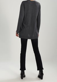 Lång grå stickad tröja med rundad halsringning, avslappnad passform och sidospjällsdetalj, kombinerad med svarta utsvängda jeans och ankelboots.