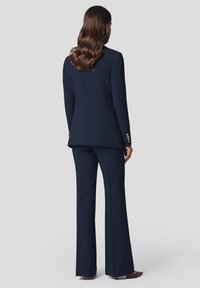 Granatowy garnitur szyty na miarę z dopasowaną marynarką i rozszerzanymi spodniami. Klasyczny design z złotymi guzikami na rękawach. Gładka tekstura materiału.