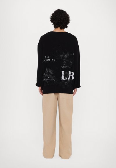 Un sweat-shirt noir oversized avec un texte graphique blanc, porté avec un pantalon large beige, affiché de dos sur un fond uni.