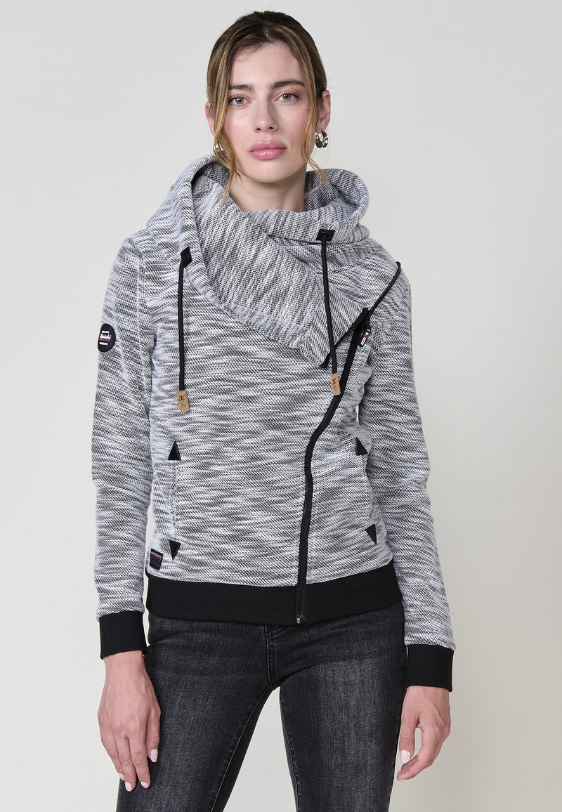 Szürke és fekete csíkos pulóver kapucnival, elöl cipzárral és állítható zsinórokkal. Fekete díszítéssel és részletekkel rendelkezik.