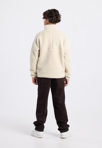 Pullover in pile color crema con colletto alto, superficie testurizzata e dettagli delle cuciture, abbinato a pantaloni in corduroy marrone scuro e sneakers bianche.