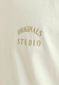 Crème-kleurig t-shirt van zacht katoen, met olijfgroene geborduurde tekst "ORIGINALS STUDIO" in een gebogen ontwerp.