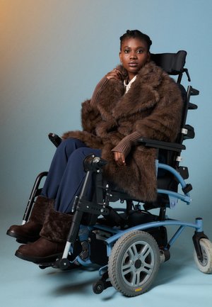 Žena v hnědém kožichu a botách sedící v světle modrém elektrickém invalidním vozíku na jednobarevném pozadí.