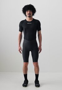 Schwarzer Radanzug mit kurzen Ärmeln, gerippter Textur, eng anliegendem Design und gepolsterten Shorts. Getragen mit schwarzen Socken und Schuhen.