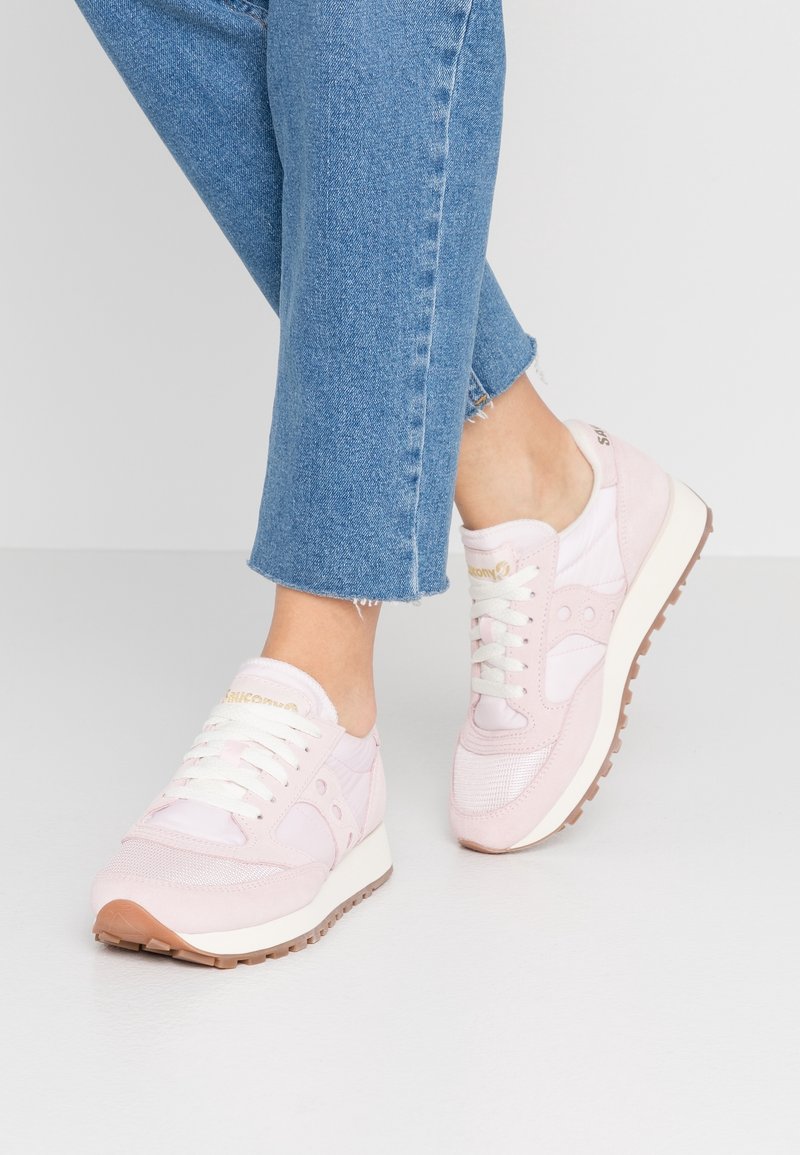 Des pieds chaussés de baskets Saucony rose pâle avec des lacets blancs et d'un jean cropped bleu, se tenant sur un sol blanc devant un fond uni.