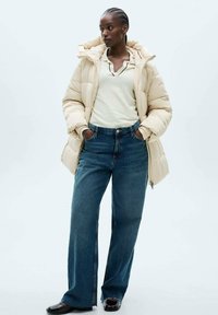 Beige pufferjack, oversized pasvorm, met een hoge kraag en ritszakken. Gecombineerd met een lichtcrème top en brede blauwe denim jeans. Zwarte schoenen.