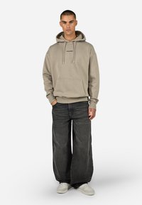 Felpa beige con tasca frontale e cappuccio regolabile, abbinata a jeans larghi scuri e sneakers bianche. Dettaglio del logo semplice sulla felpa.