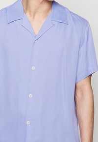 Camicetta corta a maniche corte di colore viola chiaro con colletto a camicia, dotata di cinque bottoni e una finitura in tessuto leggero e morbido.