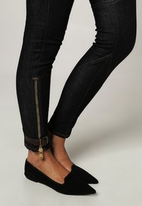 Černé džíny s úzkým střihem, se zlatým zipem a hnědým detailem u lemu. Nosit s špičatými černými balerínami.