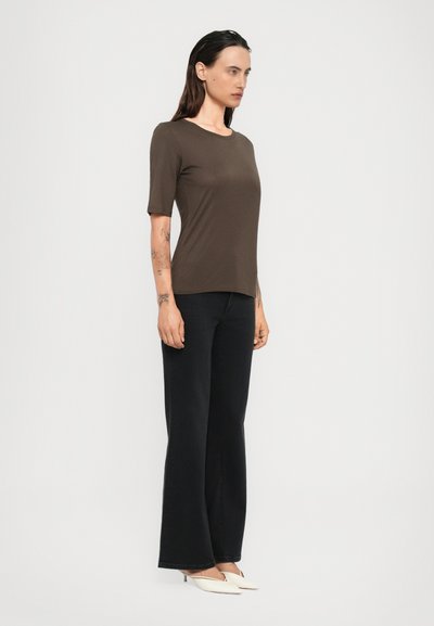 Donna in top corto marrone a maniche corte, pantaloni neri a gamba larga e tacchi a punta bianchi è in piedi rivolta verso sinistra su uno sfondo leggero e neutro.