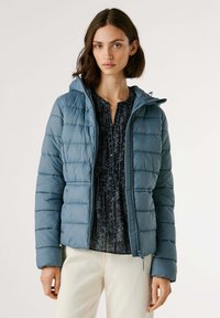 Vrouw in een blauwe gewatteerde jas met capuchon, over een donker gemarmerde blouse en lichtgekleurde broek, staande tegen een effen achtergrond.